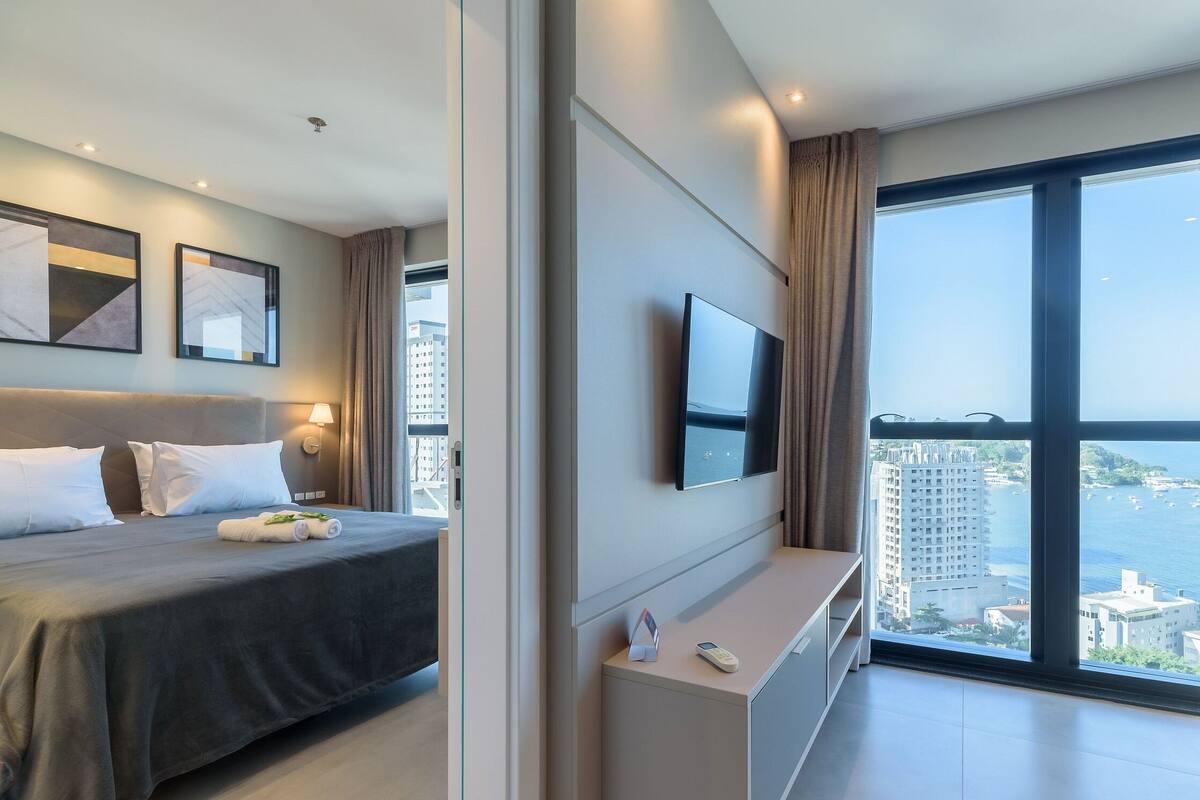 A modern hotel room with a bed, a TV mounted on the wall, a large window with a city view, and a small table with a remote control.