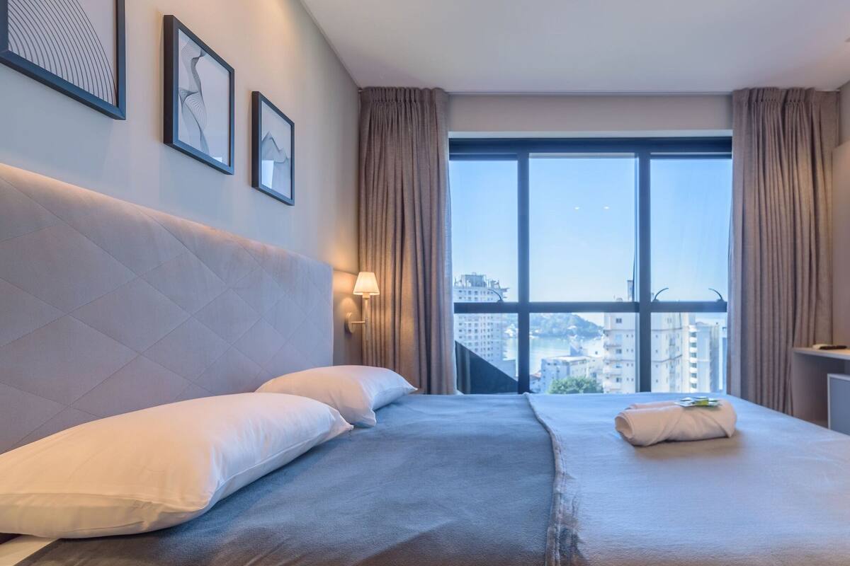 A hotel room with a large bed, a TV, a window with a city view, and framed artwork on the wall.