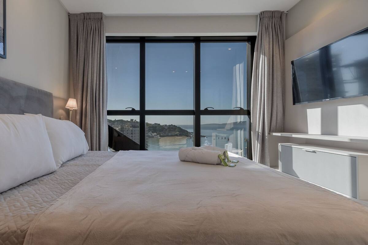 A hotel room with a large bed, a TV mounted on the wall, and a view of a cityscape through a large window.