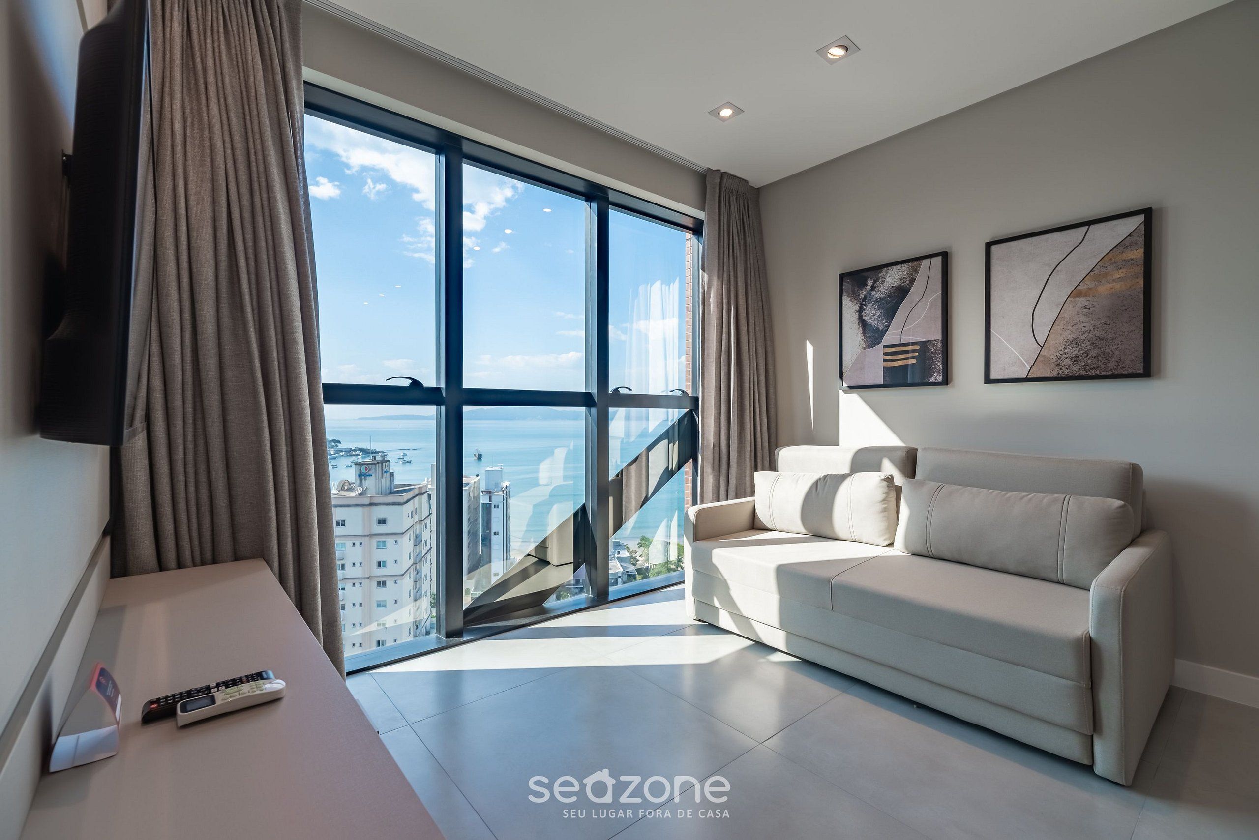 Una sala de estar moderna con un sofá, un televisor montado en la pared y un ventanal con vistas a edificios y al mar.
