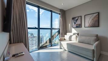 Una sala de estar moderna con un sofá, un televisor montado en la pared y un ventanal con vistas a edificios y al mar.