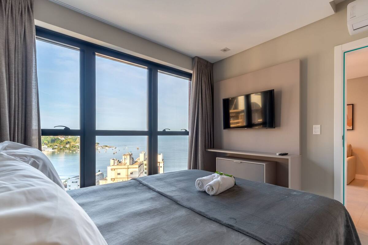 A hotel room with a large window, a bed, a TV, and a view of the sea.