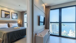 Une chambre d’hôtel moderne équipée d’un lit, d’une télévision à écran plat, d’un petit bureau et d’une grande fenêtre donnant sur la ville.
