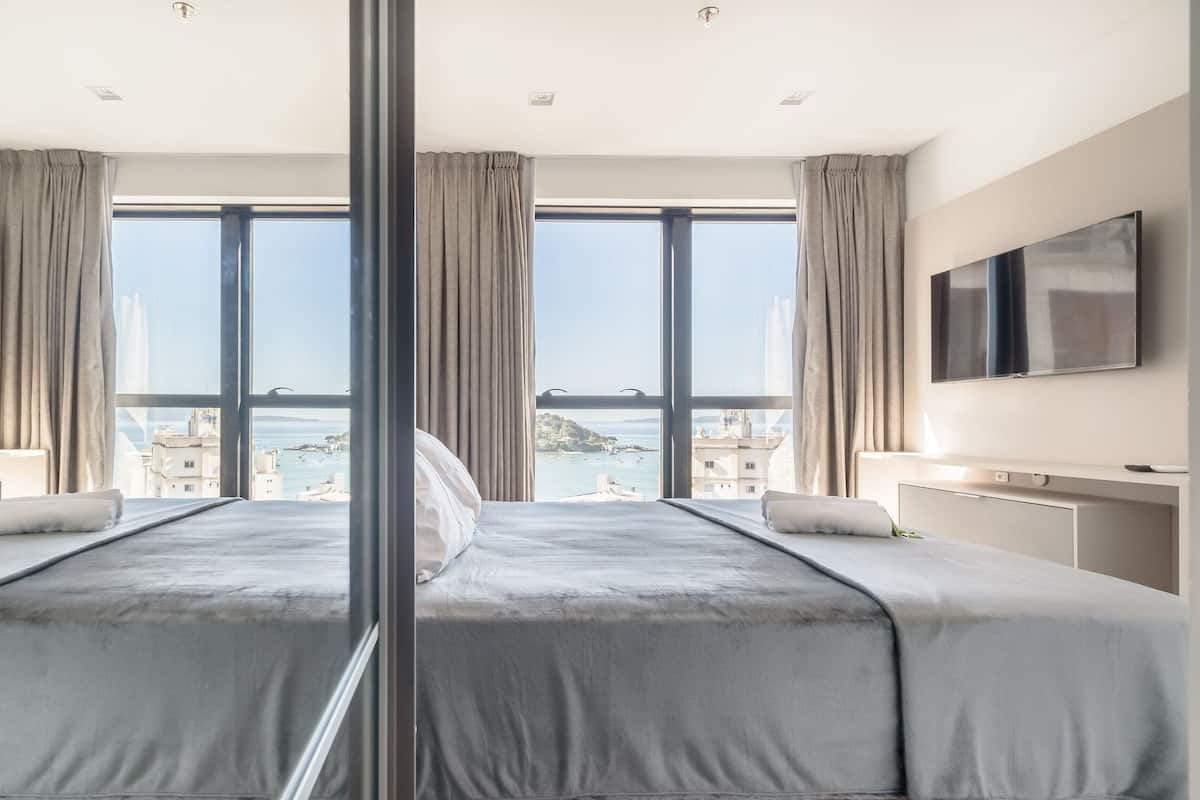 A hotel room with a large bed, two TV screens, and a view of a coastal city.
