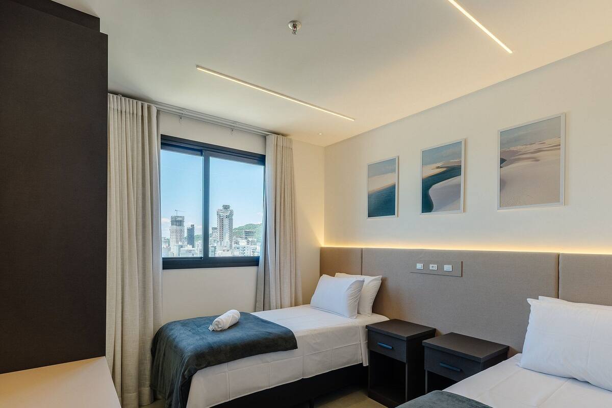 A hotel room with two beds, a large window with a city view, and three framed pictures on the wall.