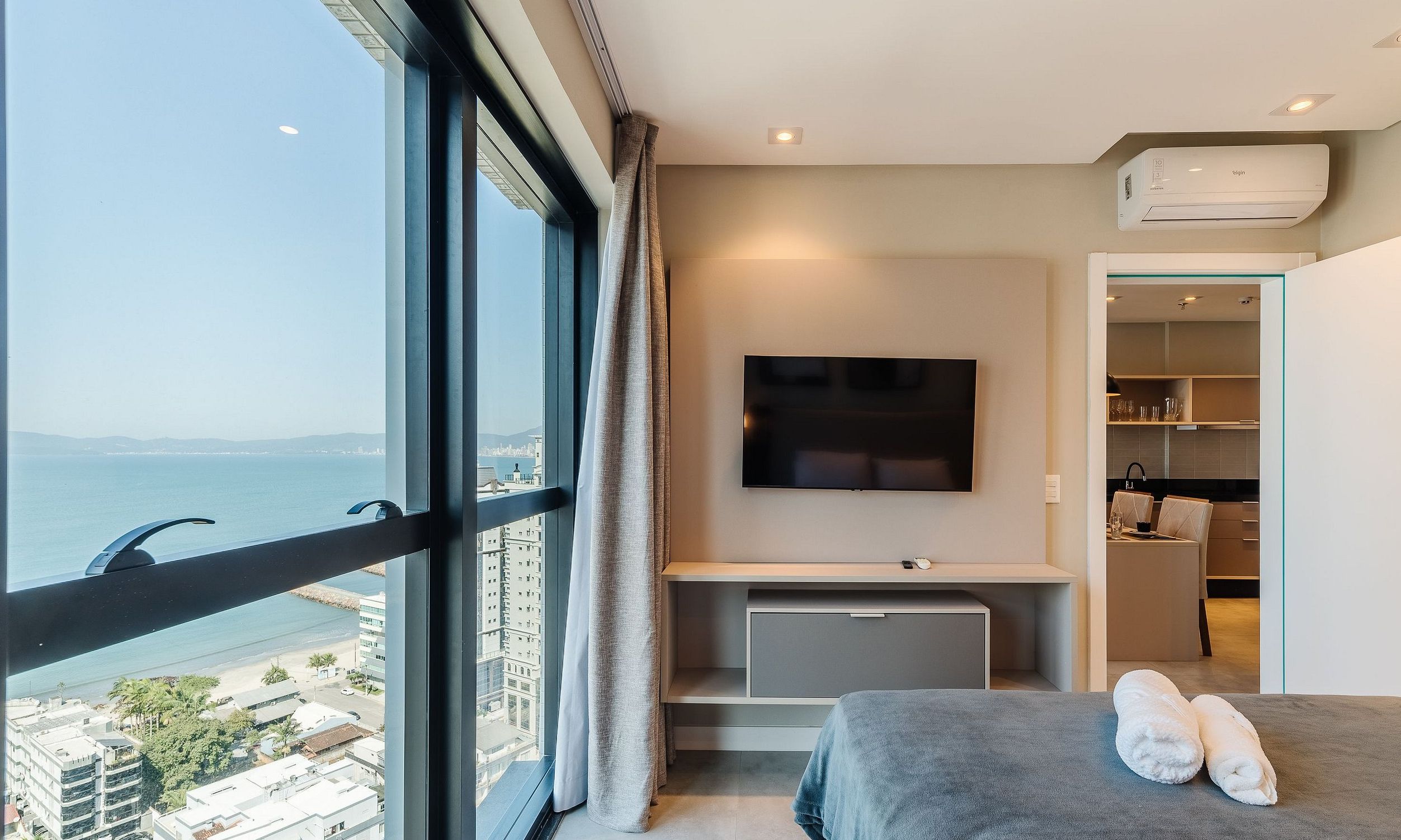 A modern hotel room with a large window, a flat-screen TV mounted on the wall, a small kitchenette, and a bed with two towels.
