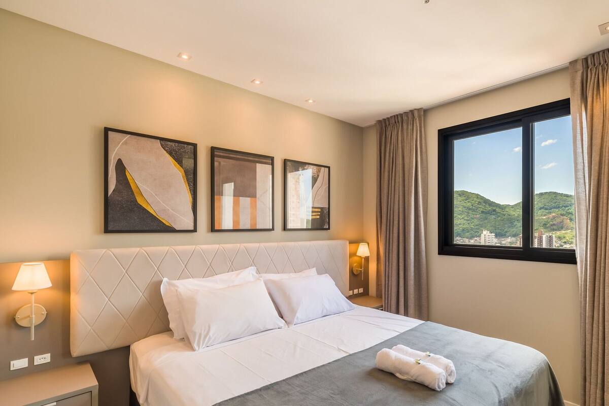 A hotel room with a bed, two wall-mounted lamps, a window with curtains, and three framed artworks on the wall.