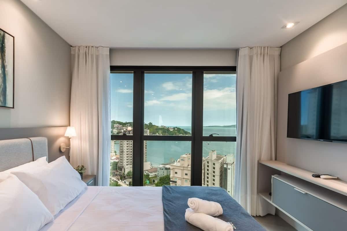 A hotel room with a bed, a TV, a window with a city view, and a painting on the wall.