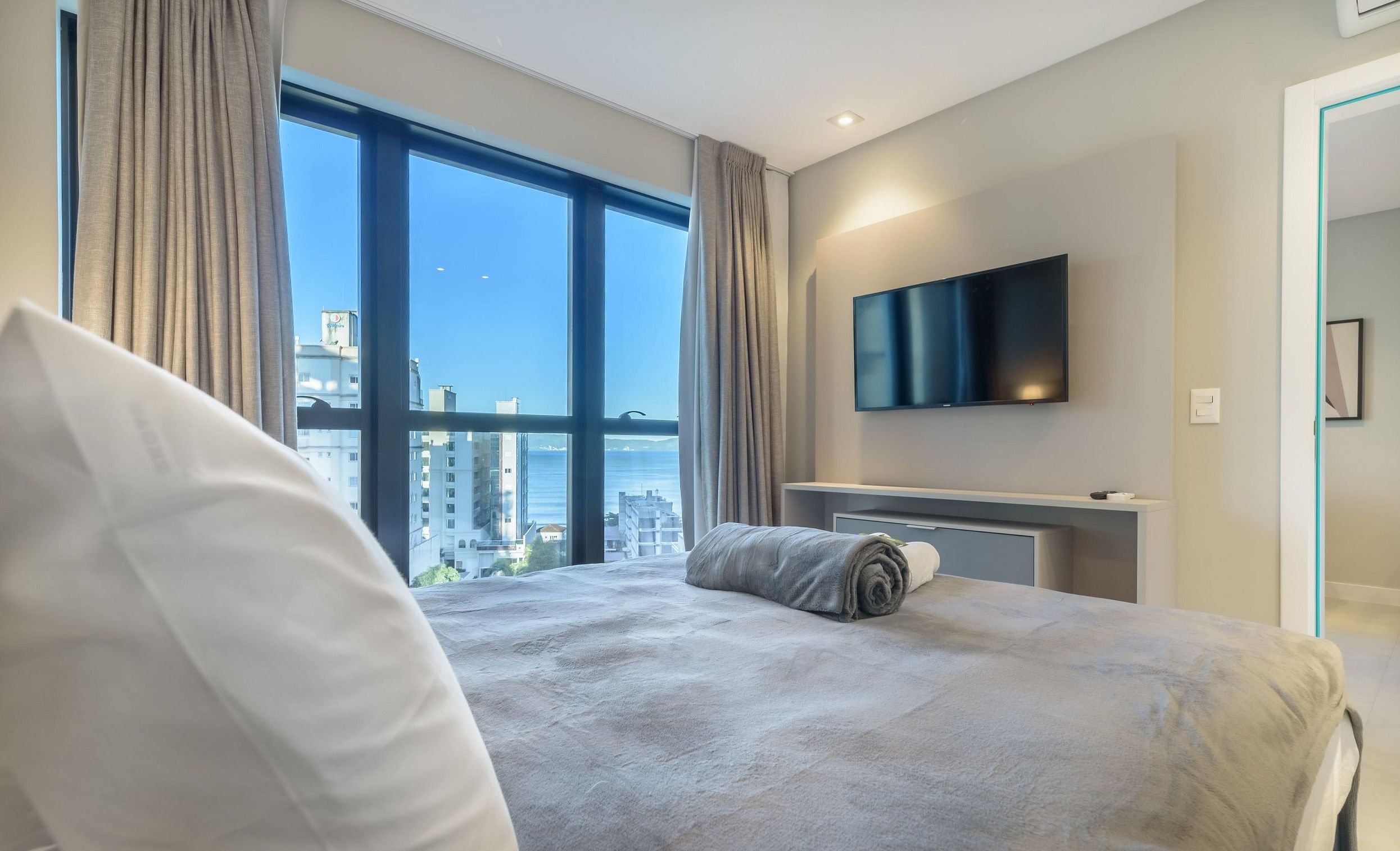 A hotel room with a large bed, a flat-screen TV mounted on the wall, and a view of the city through large windows.