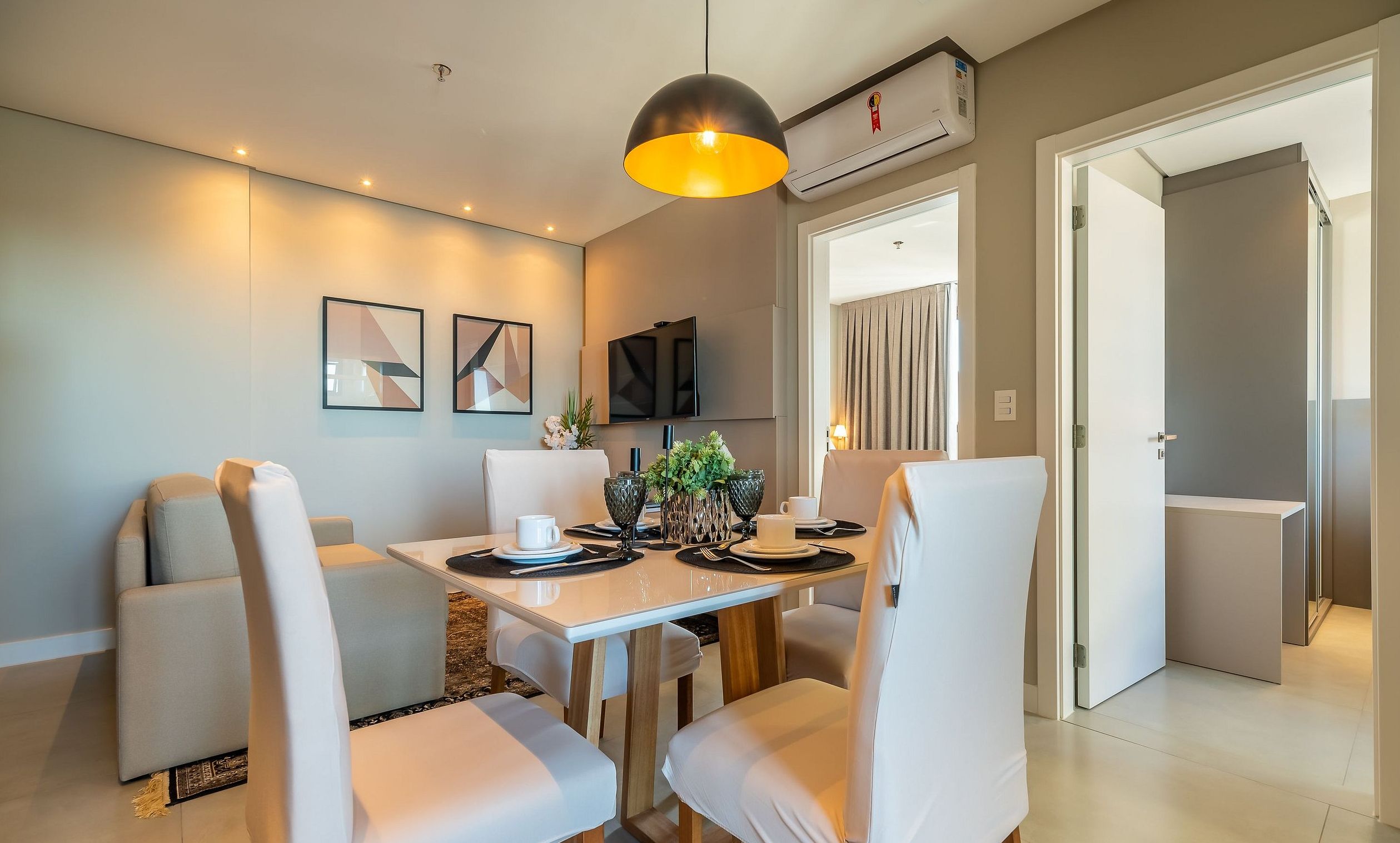 Un comedor moderno con una mesa y sillas blancas, una lámpara colgante y un televisor de pantalla plana montado en la pared.