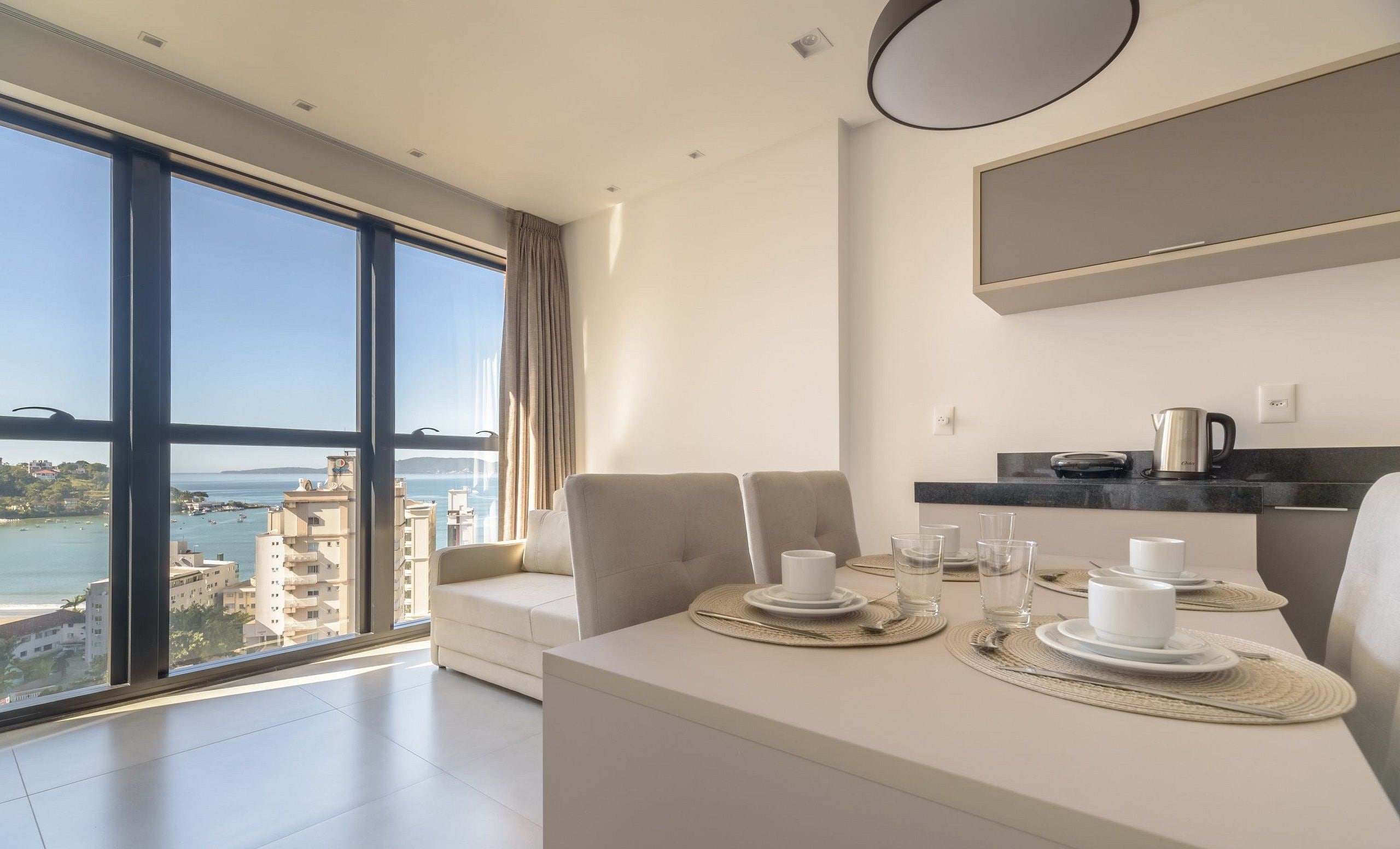 A modern hotel room with a dining area, a large window with a view of the sea and buildings, and a minimalist design.