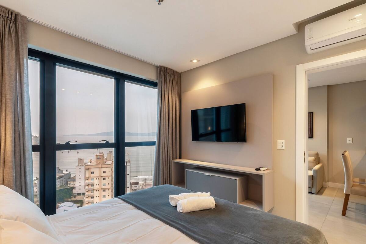 A hotel room with a large window, a flat-screen TV, a bed with white linens, and a grey bedside table.