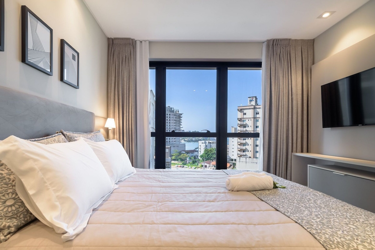Une chambre d’hôtel avec un grand lit, une télévision, une fenêtre donnant sur la ville et des œuvres d’art encadrées accrochées au mur.