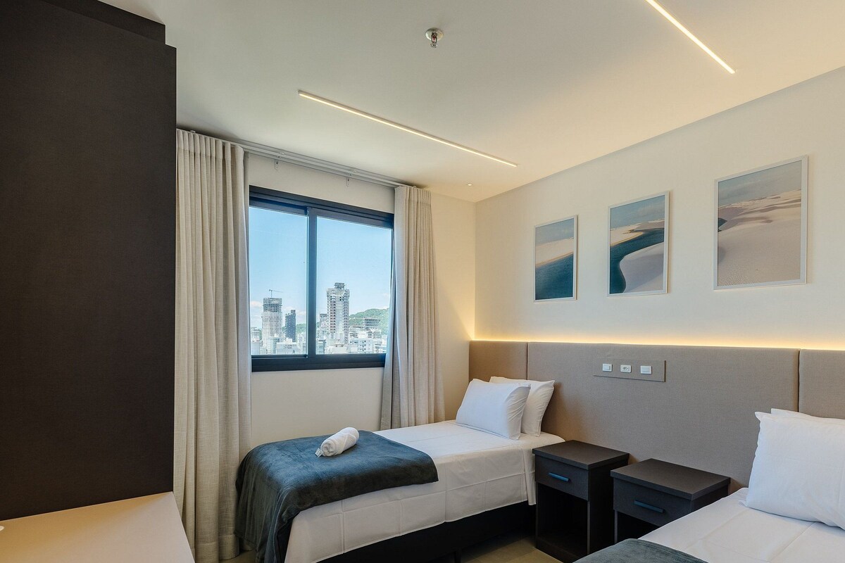 Habitación de hotel con dos camas, un ventanal con vistas a la ciudad y tres cuadros en la pared.