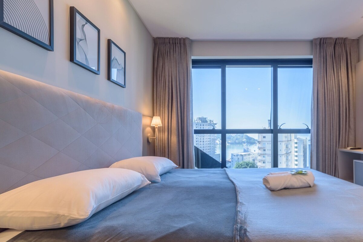 Une chambre d’hôtel avec un grand lit, une télévision, une fenêtre donnant sur la ville et des œuvres d’art encadrées accrochées au mur.