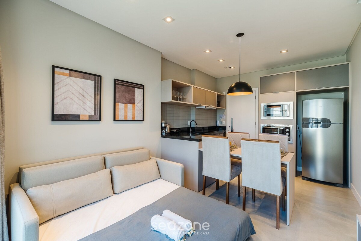 A modern hotel room with a sofa, a dining table with chairs, a kitchen area with a microwave and refrigerator, and artwork on the walls.