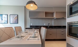 A modern kitchen with a dining area, featuring a built-in oven, a large pendant light, and a dining table set for four. - MHFs Apartamentos com Vista Mar Itapema (Itapema)