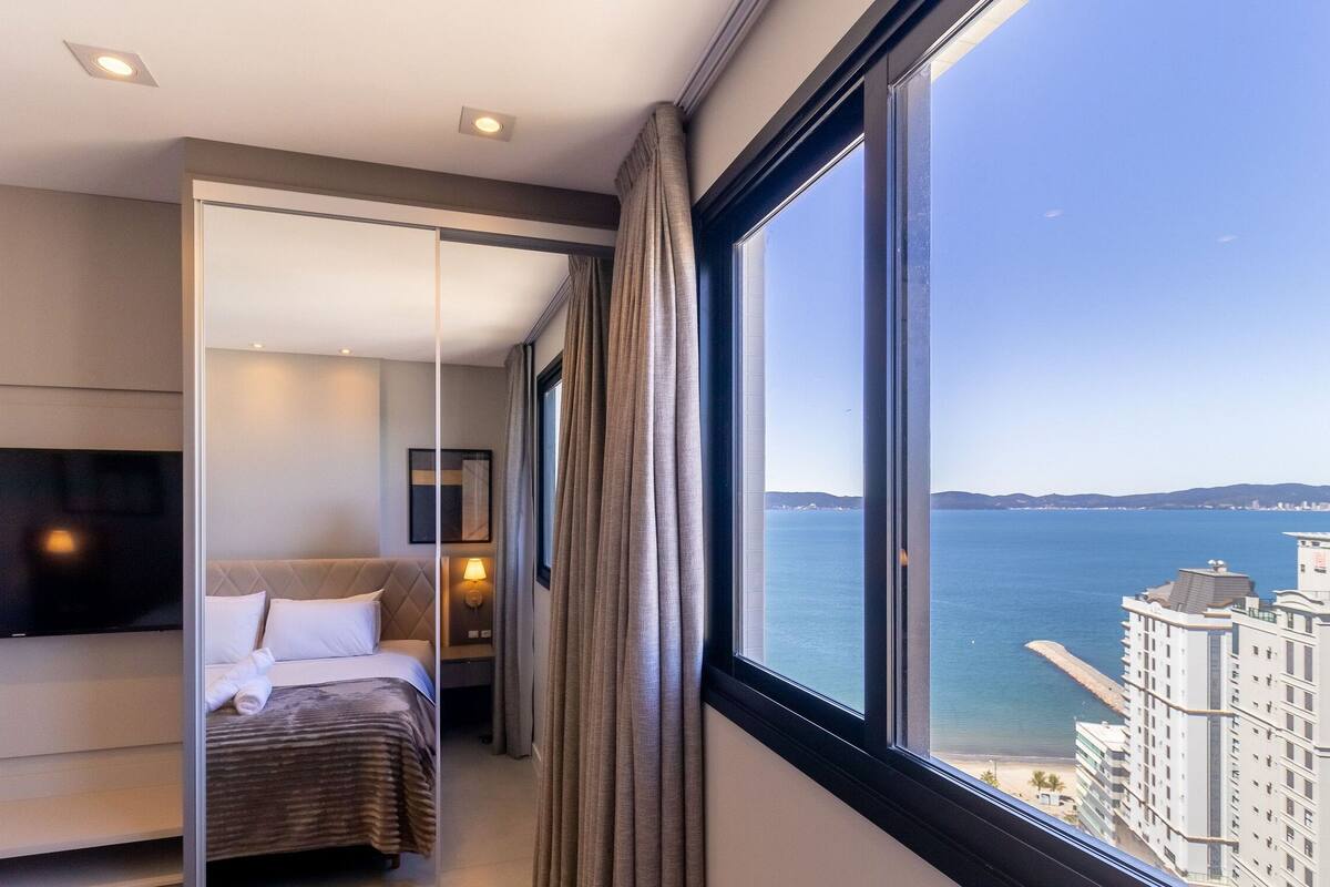 A modern hotel room with a bed, a TV, a bedside table with a lamp, and a large window offering a view of the sea and buildings.