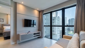 A modern living room with a flat-screen TV, a sofa, and a large window offering a city view. - MHFs Apartamentos com Vista Mar Itapema (Itapema)