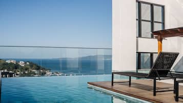 A modern infinity pool with a clear view of the sea and boats, a wooden deck, and a lounge chair.