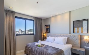 A hotel room with a bed, a nightstand, a lamp, a window with curtains, and a city view. - MHFs Apartamentos com Vista Mar Itapema (Itapema)