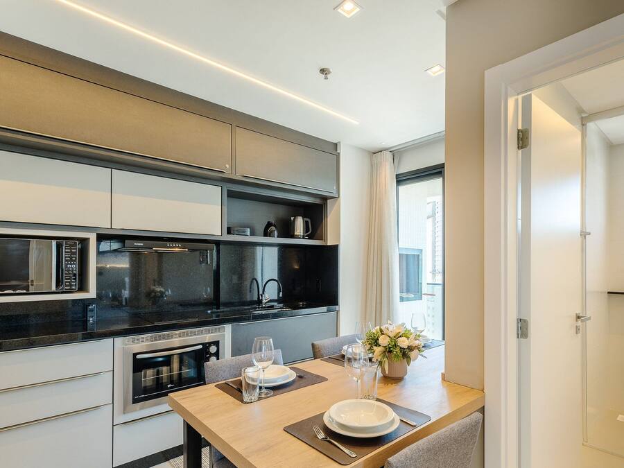 Una cocina moderna con electrodomésticos integrados, una zona de comedor con una mesa lista para dos personas y vistas de la ciudad a través de amplias ventanas.