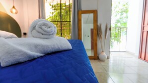 A bed with a blue quilt and a rolled white towel, a wooden door, and a view of greenery through a window. - Friendly House (Santa Tecla)
