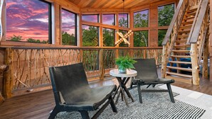 A wooden cabin with a large window overlooking a sunset, two chairs, a small table with a plant, and a wooden staircase. - NEW Canopy Crest Cabin~Indoor Pool~Hot Tub~Game Room (Sevierville)
