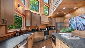A modern kitchen with wooden cabinets, stainless steel appliances, and a large window overlooking greenery. - NEW Canopy Crest Cabin~Indoor Pool~Hot Tub~Game Room (Sevierville)