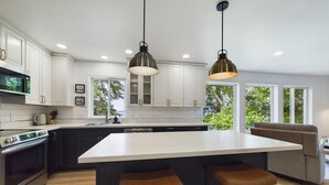 A modern kitchen with white cabinets, a large island with bar stools, and pendant lighting. - Lena Beach Oceanview Suite (Juneau)