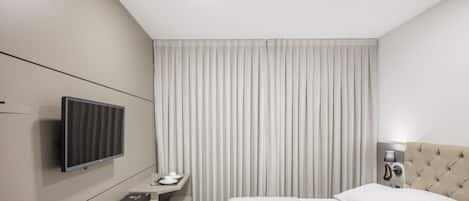 Una habitación de hotel moderna con una cama, un escritorio con televisor de pantalla plana, una mesita de noche y un ventanal con cortinas.