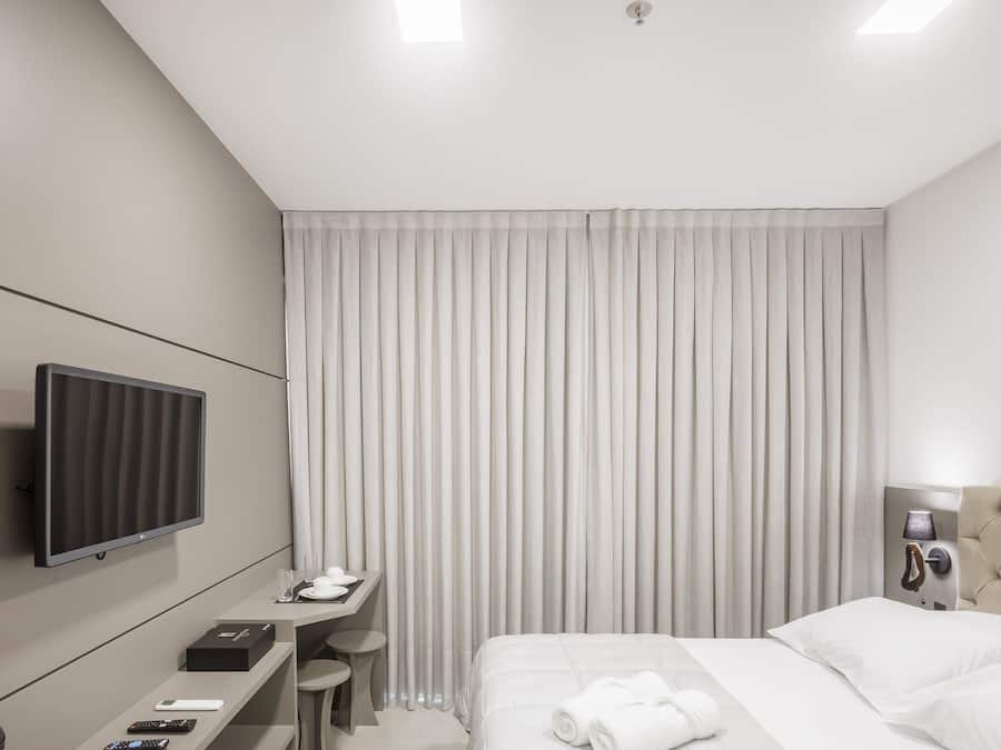 Una habitación de hotel moderna con una cama, un escritorio con televisor de pantalla plana, una mesita de noche y un ventanal con cortinas.