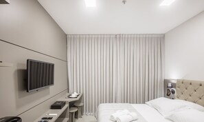 Una habitación de hotel moderna con una cama, un escritorio con televisor de pantalla plana, una mesita de noche y un ventanal con cortinas.