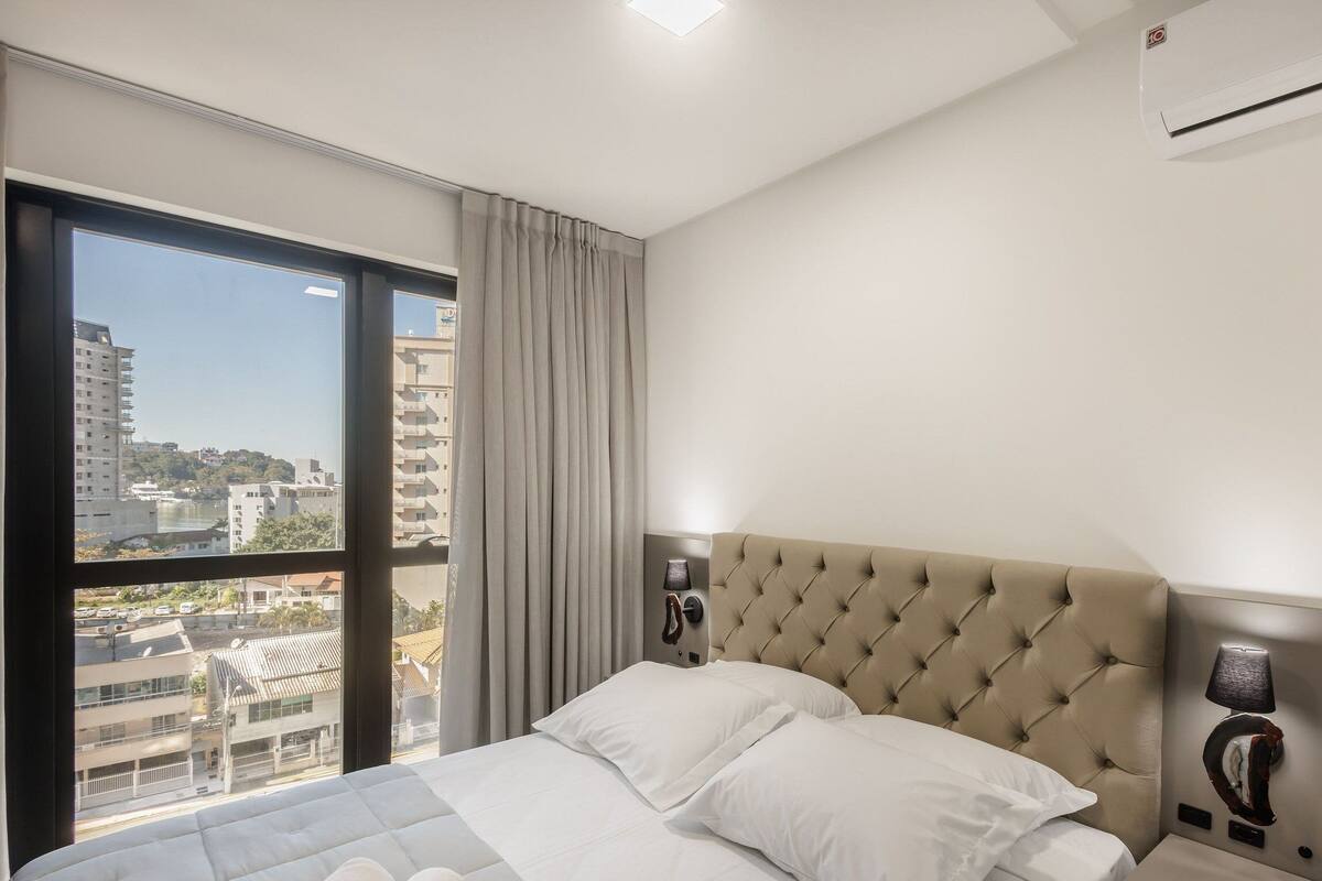 A hotel room with a large window, a tufted headboard, a bedside lamp, and an air conditioning unit.