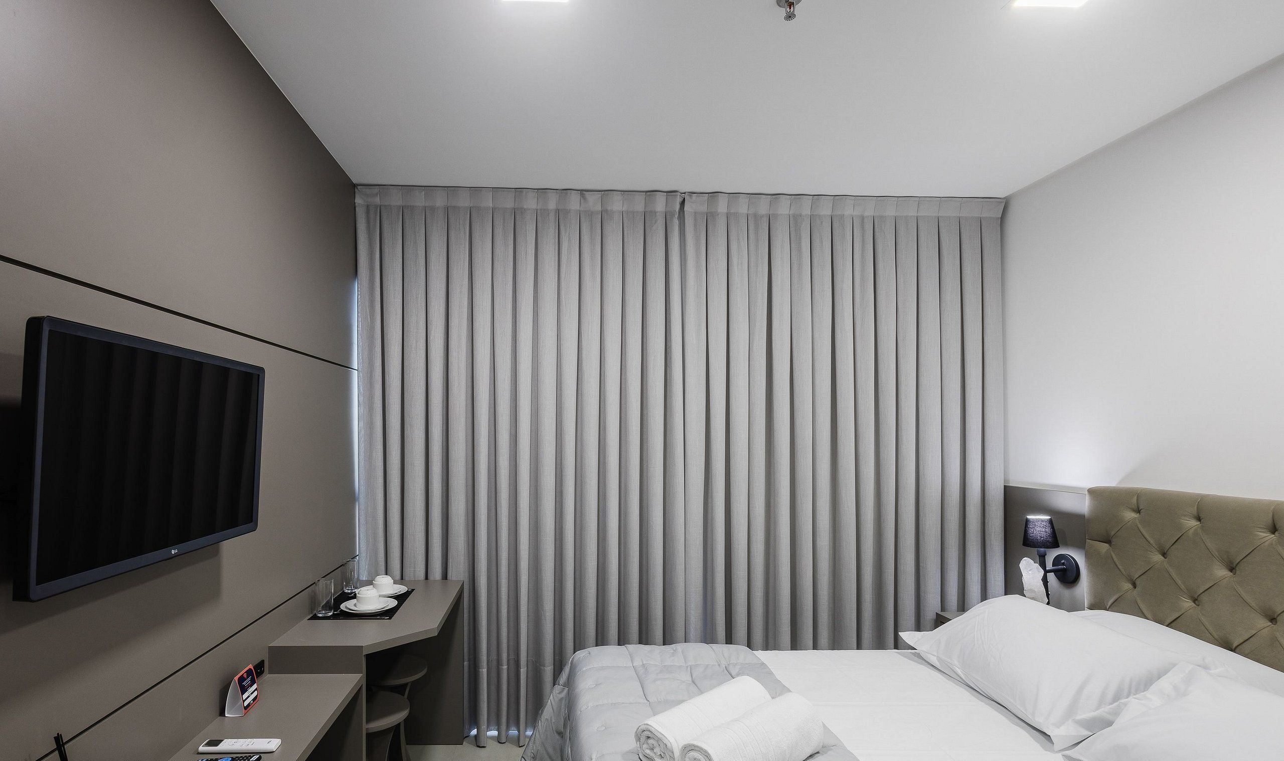A hotel room with a bed, a desk with a computer, a TV, and a window with curtains.