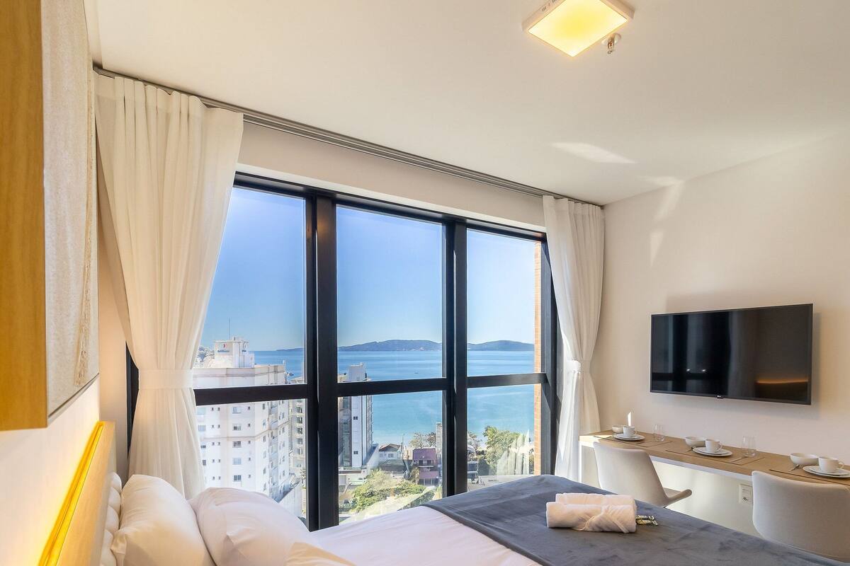 A hotel room with a bed, a desk, a TV, and a large window with a view of the sea.