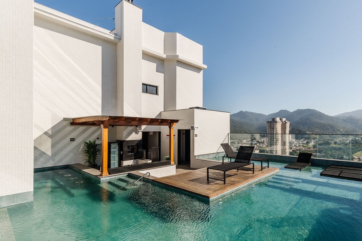 Un edificio moderno con piscina infinita en la azotea, mobiliario exterior y vistas a montañas.