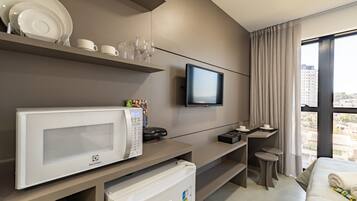 Una habitación de hotel moderna con televisor de pantalla plana, microondas, refrigerador pequeño, escritorio con silla y un amplio ventanal con vista a la ciudad.