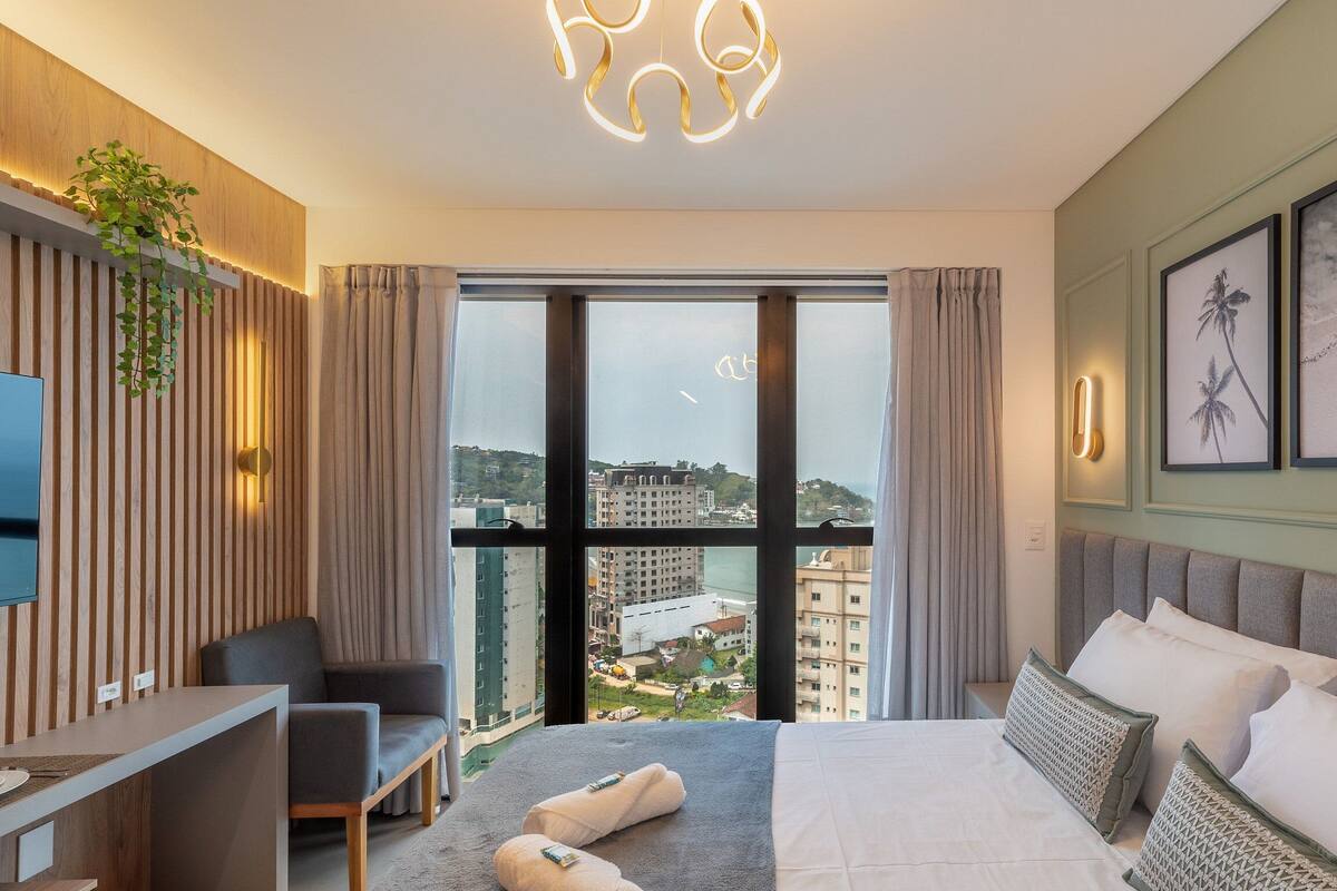 A modern hotel room with a large bed, a desk, a chair, and a view of the cityscape through floor-to-ceiling windows.