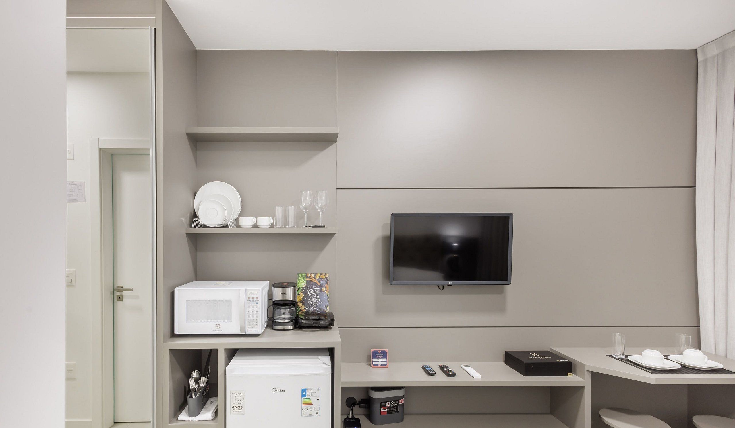 Une chambre d’hôtel moderne équipée d’un téléviseur à écran plat fixé au mur, d’un four à micro-ondes, d’une machine à café et d’une petite table sur laquelle se trouvent une télécommande et un téléphone intelligent.