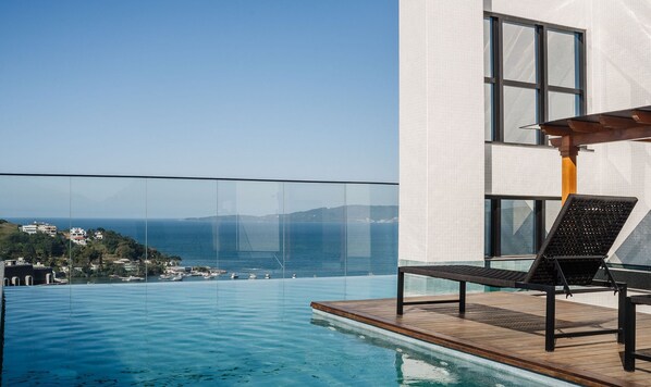 A modern infinity pool with a clear view of the sea and boats, a wooden deck, and a lounge chair. - MHFA Studios em Prédio Luxuoso Vista Mar (Itapema)