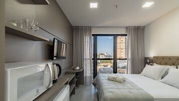 A hotel room with a bed, a TV, a kettle, and a view of buildings through the window.