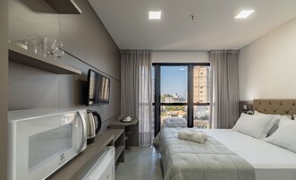 A hotel room with a bed, a TV, a kettle, and a view of buildings through the window.