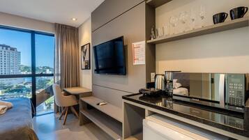 A modern hotel room with a bed, a desk, a flat-screen TV, a minibar, and a large window with a city view.