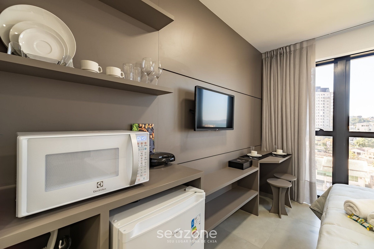 Una habitación de hotel moderna con televisor de pantalla plana, microondas, refrigerador pequeño, escritorio con silla y un amplio ventanal con vista a la ciudad.