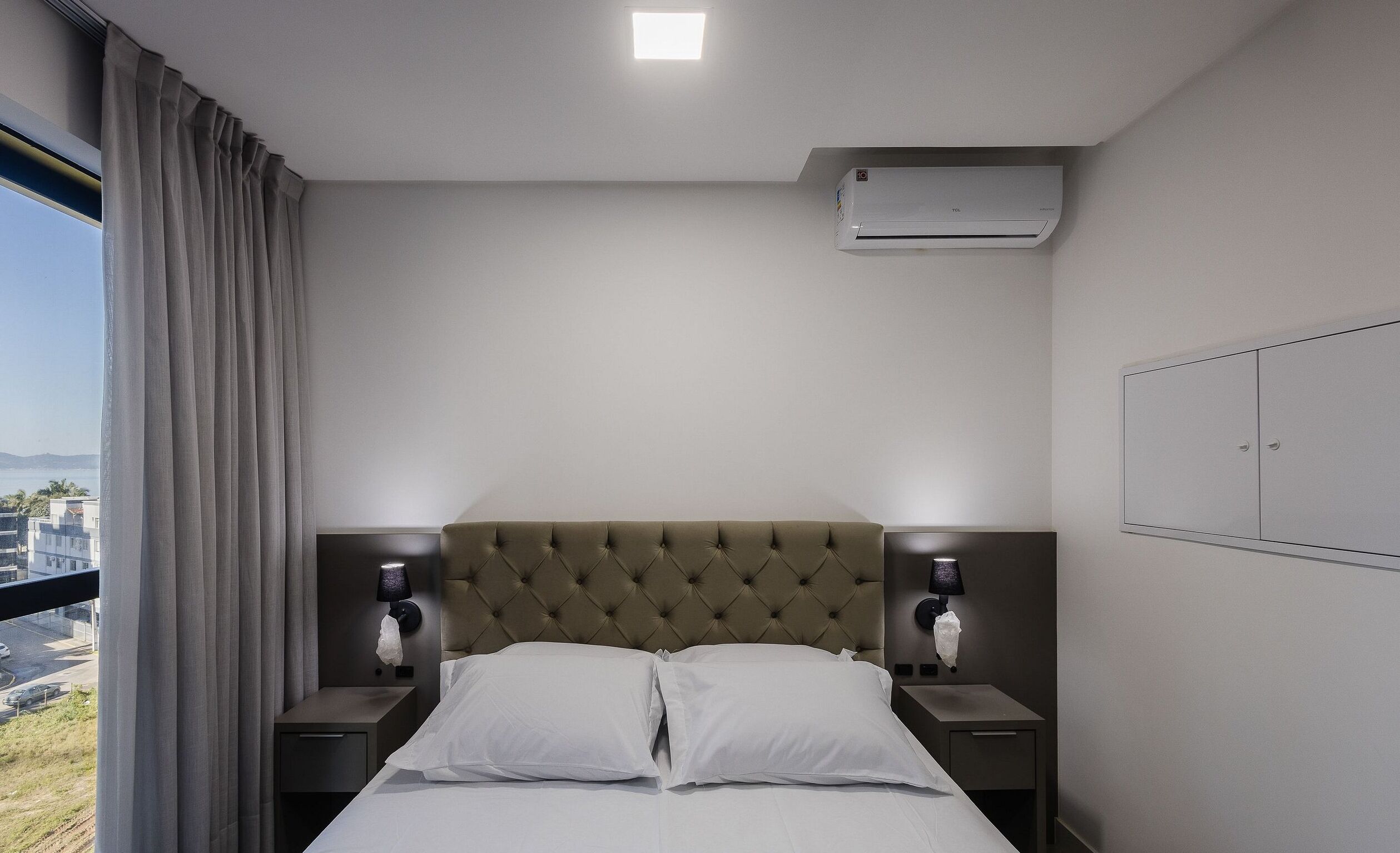Habitación de hotel con cama, dos mesitas de noche, aire acondicionado de pared y vistas a la ciudad a través de la ventana.