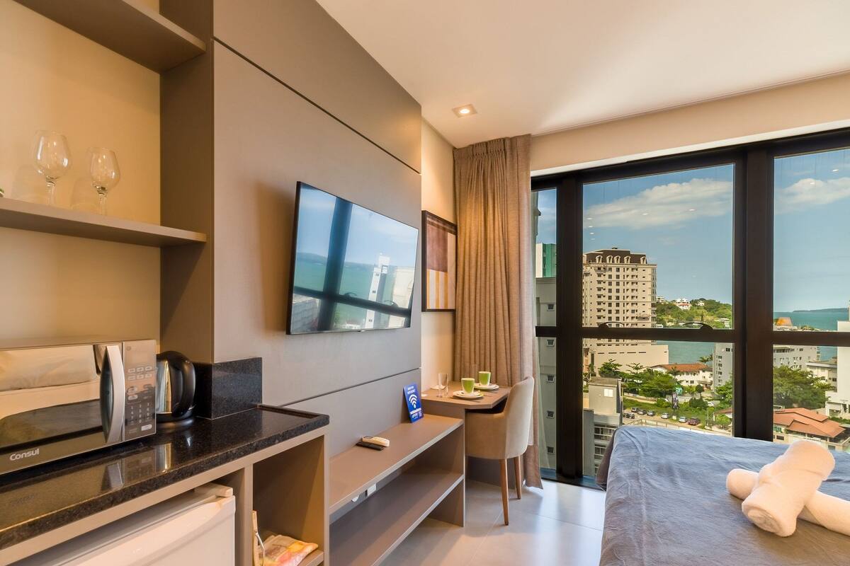 A modern hotel room with a bed, a desk, a flat-screen TV, a minibar, and a large window with a city view.