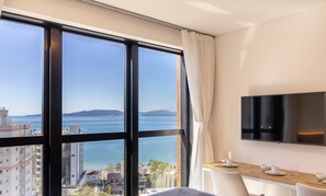 Habitación de hotel con un amplio ventanal que ofrece vistas al mar y a edificios, un televisor de pantalla plana montado en la pared y una pequeña zona de comedor con mesa y sillas.