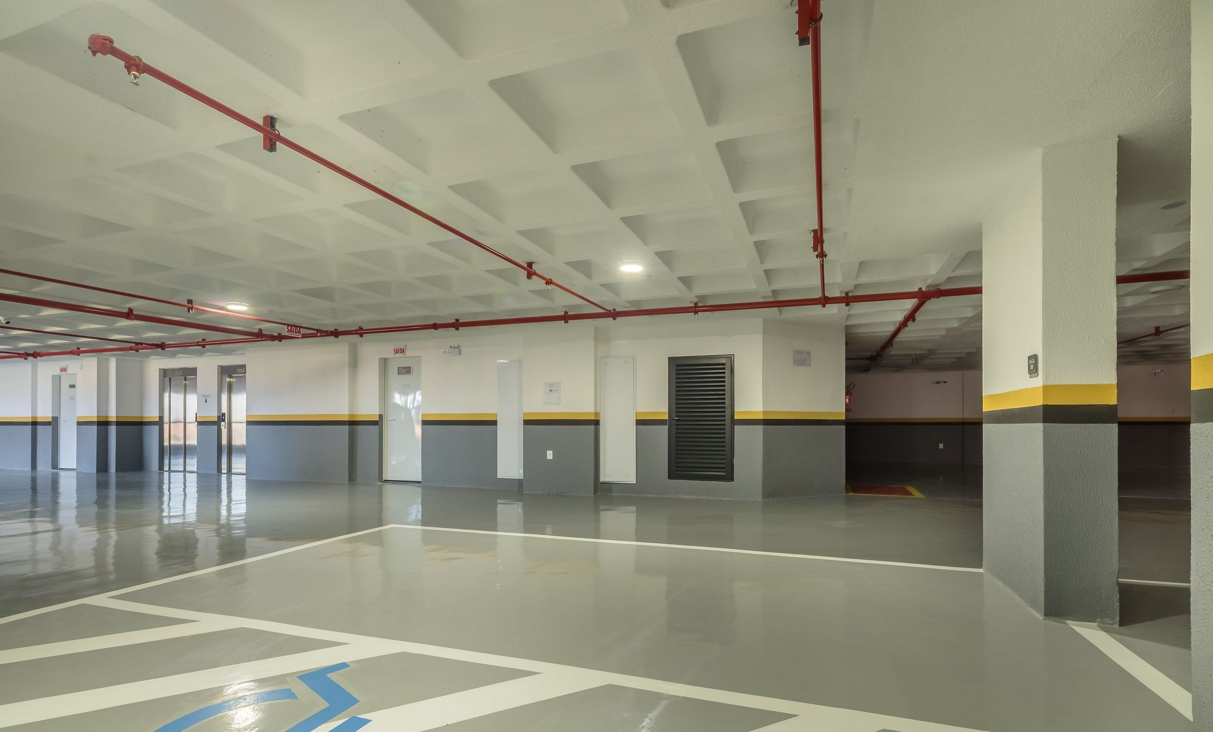 Un amplio estacionamiento interior con líneas de techo definidas, tuberías rojas y un camino accesible para sillas de ruedas.
