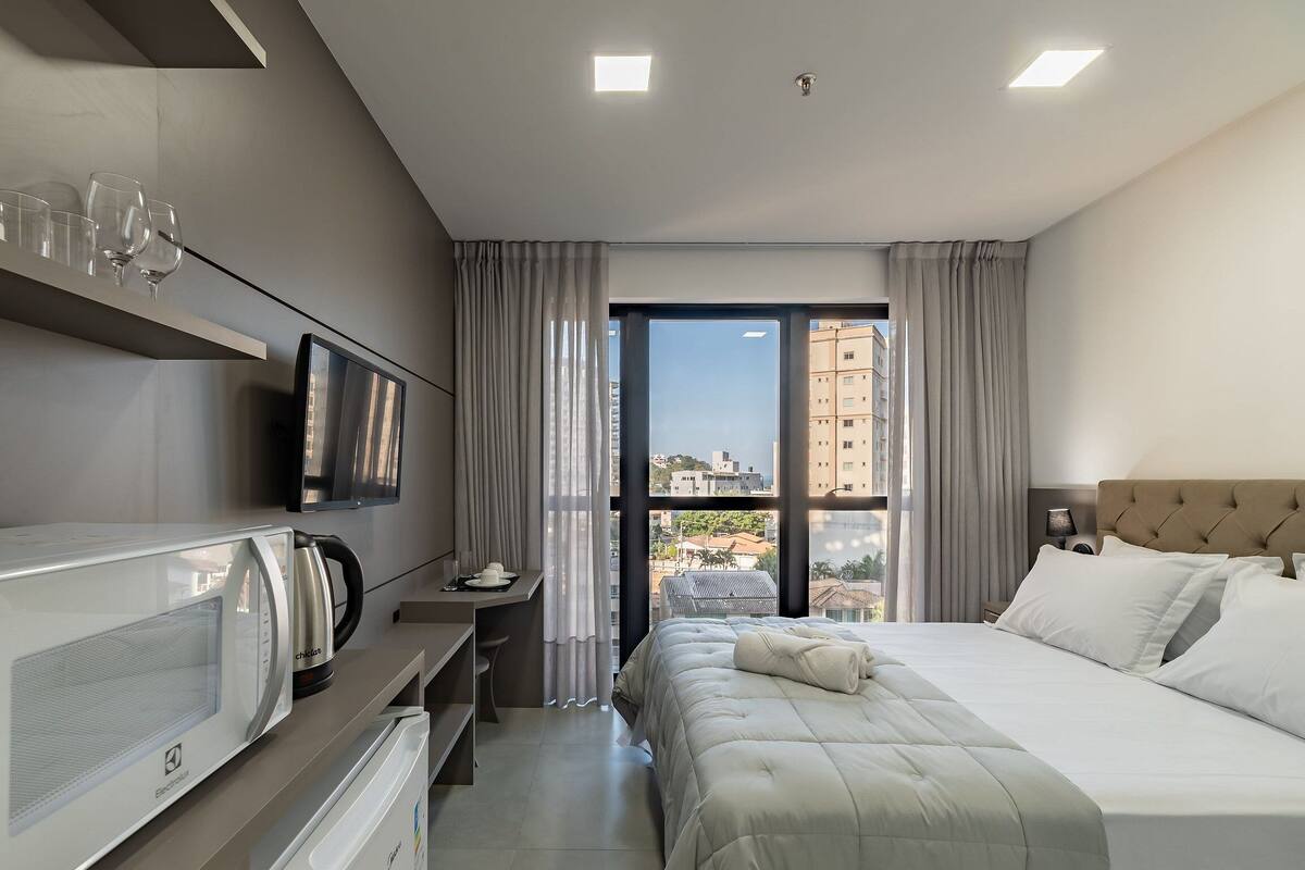 A hotel room with a bed, a desk, a TV, and a view of buildings.